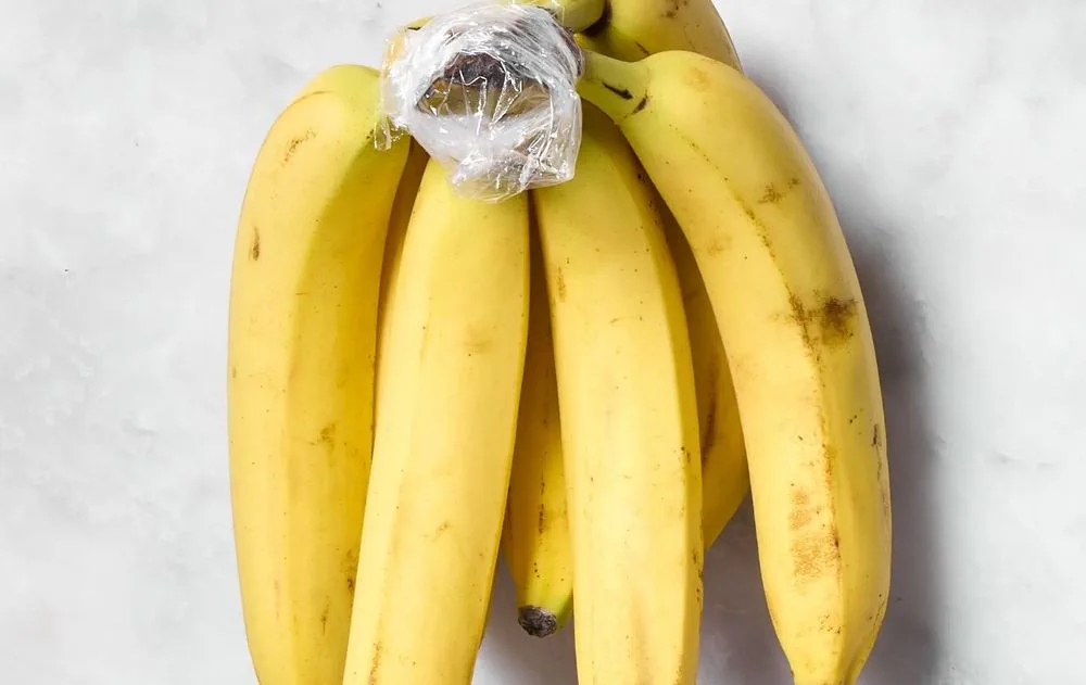 Un racimo de bananas amarillas sobre una superficie blanca tiene plástico envolviendo el cabo superior.