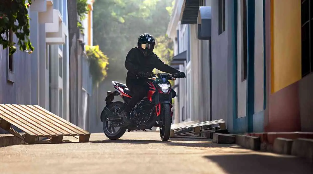Una persona con casco y campera negra conduce una moto por una calle angosta entre edificios.