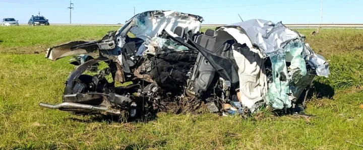 Un auto blanco totalmente destrozado y deformado yace sobre el pasto al costado de una ruta.