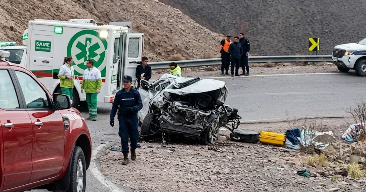 Un auto blanco destrozado al costado de una ruta cerca de una ambulancia y personal policial.