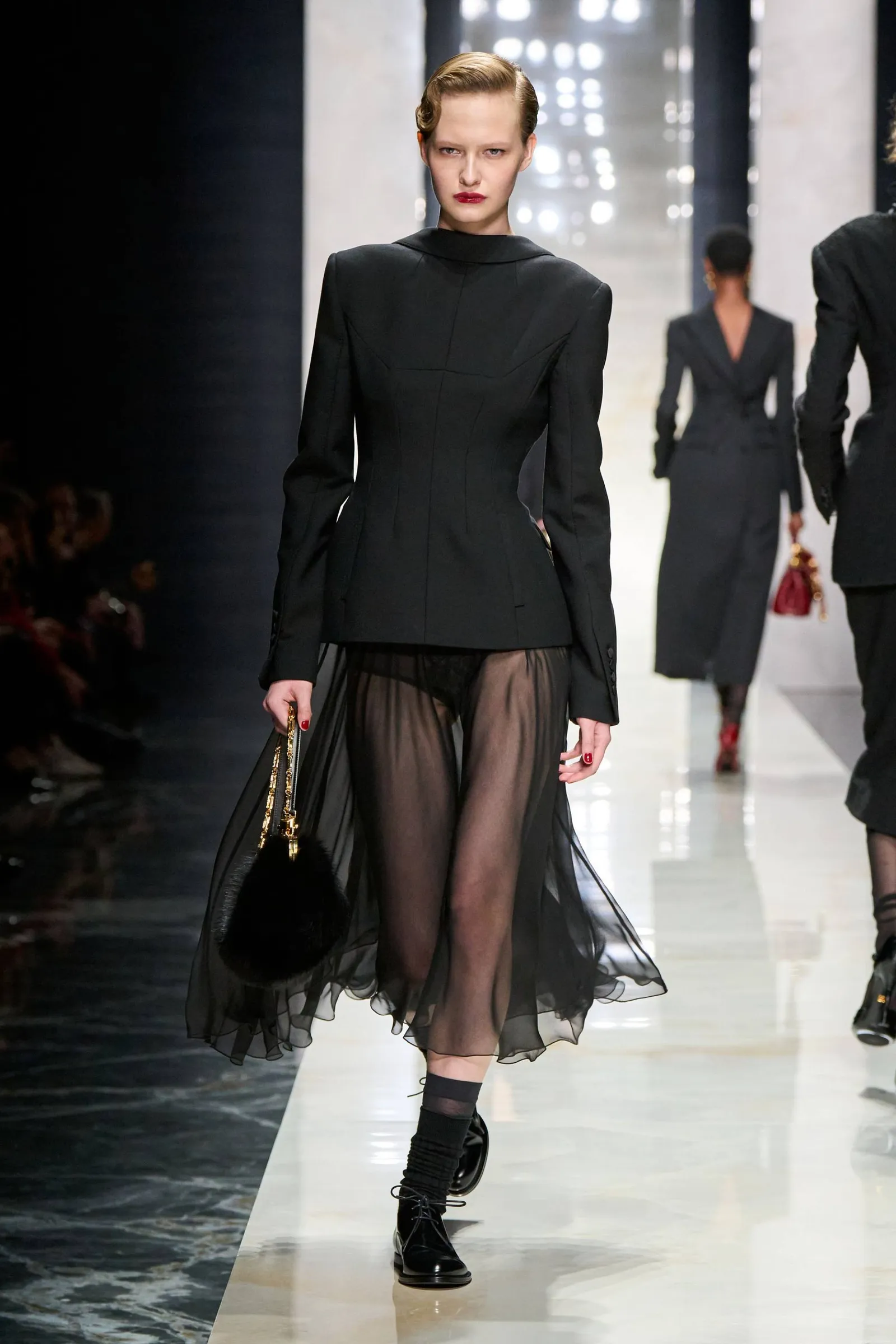 Modelo desfilando en una pasarela con saco negro entallado y pollera negra transparente.