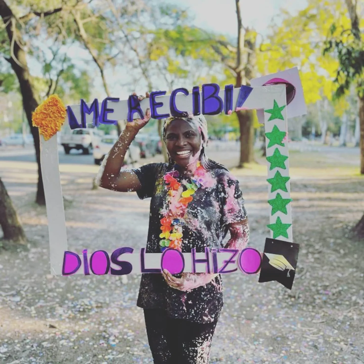 Una joven sostiene sonriente un marco de egresada con las frases Me recibí y Dios lo hizo en un parque arbolado.