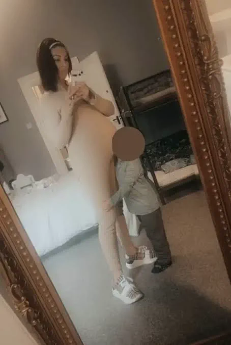 Una mujer embarazada se saca una foto frente al espejo mientras un nene la abraza de las piernas.