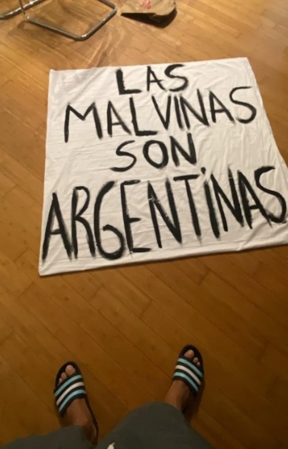 Persona en ojotas parada frente a un cartel de tela en el piso que dice "LAS MALVINAS SON ARGENTINAS".