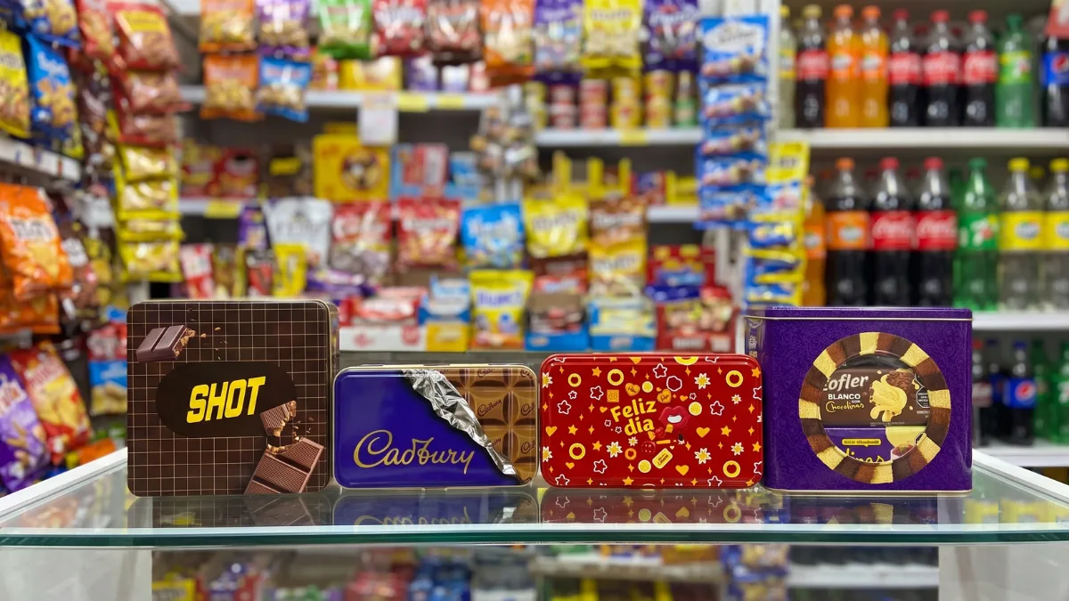 Cuatro latas metálicas de golosinas apoyadas en fila sobre un estante de vidrio dentro de un comercio.