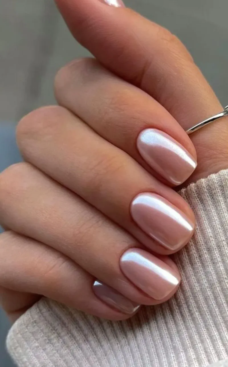 Una mano con uñas cortas y redondeadas pintadas en un tono nacarado claro sobre una prenda de punto beige.