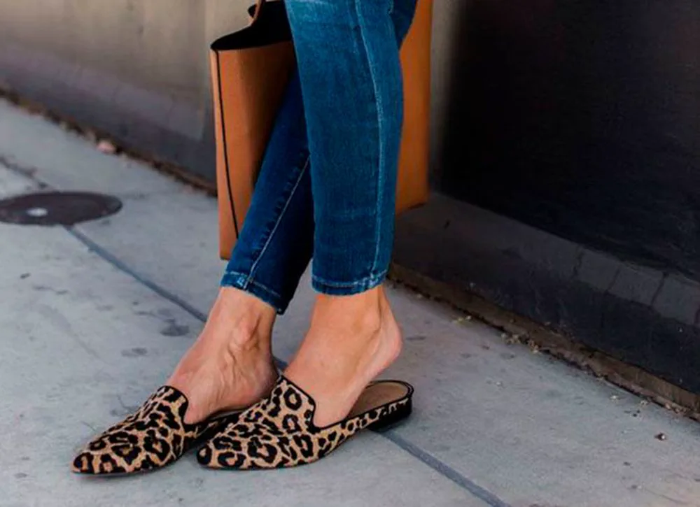 Pies de una persona con jeans y zapatos planos tipo mules con estampado animal de leopardo sobre la vereda.