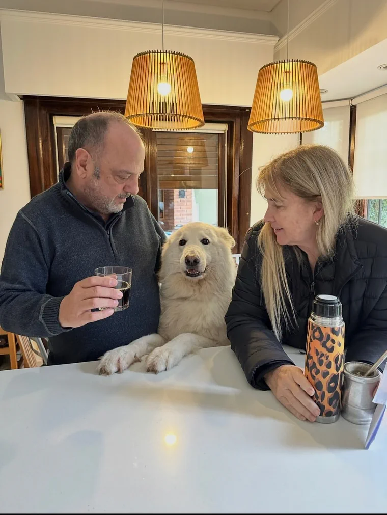 Baby Etchecopar y una mujer sentados a una mesa junto a un perro blanco, con un vaso y un termo con mate.