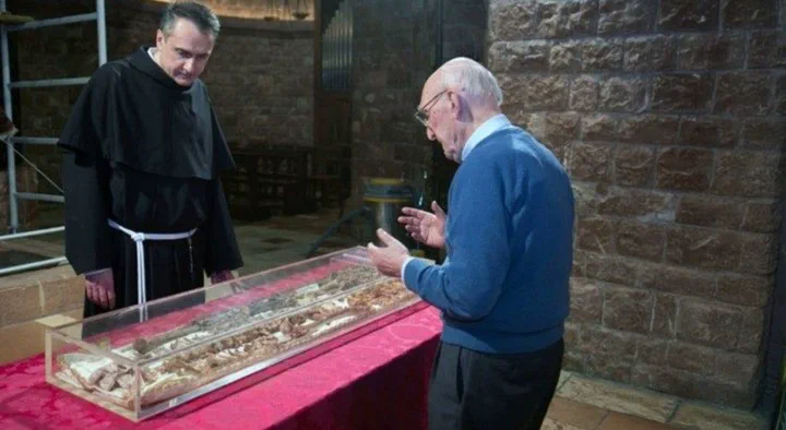 Un fraile franciscano y un hombre con anteojos observan restos óseos exhibidos dentro de una urna de acrílico.