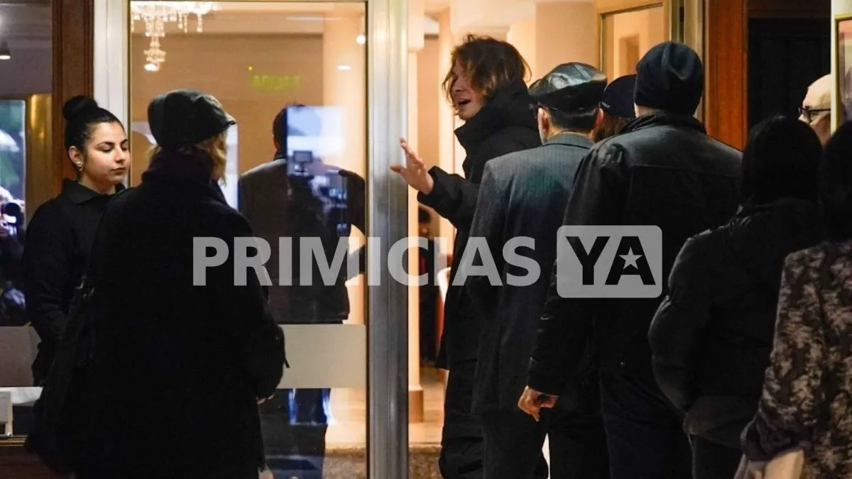 Un grupo de personas abrigadas conversando en el ingreso a un edificio vidriado de noche.