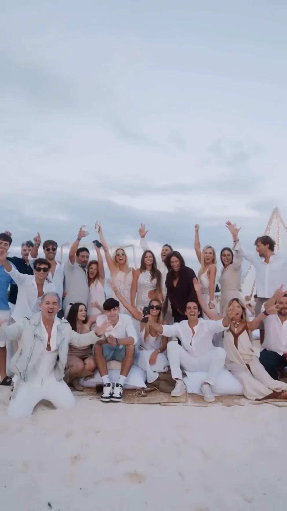 Grupo numeroso de personas vestidas de blanco posa y festeja sobre la arena de una playa.