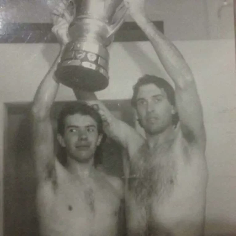 Dos hombres con el torso desnudo sostienen un trofeo en alto.