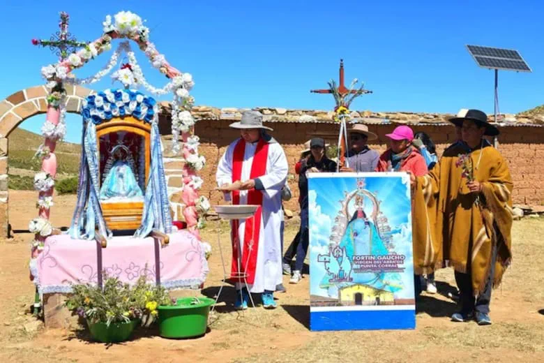 Un sacerdote y varias personas se encuentran frente a una ermita con una imagen religiosa al aire libre.