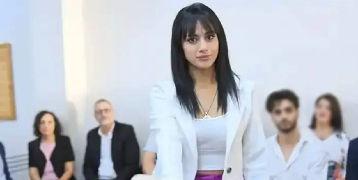 Una joven con blazer blanco mira hacia adelante en una sala con gente sentada detrás.