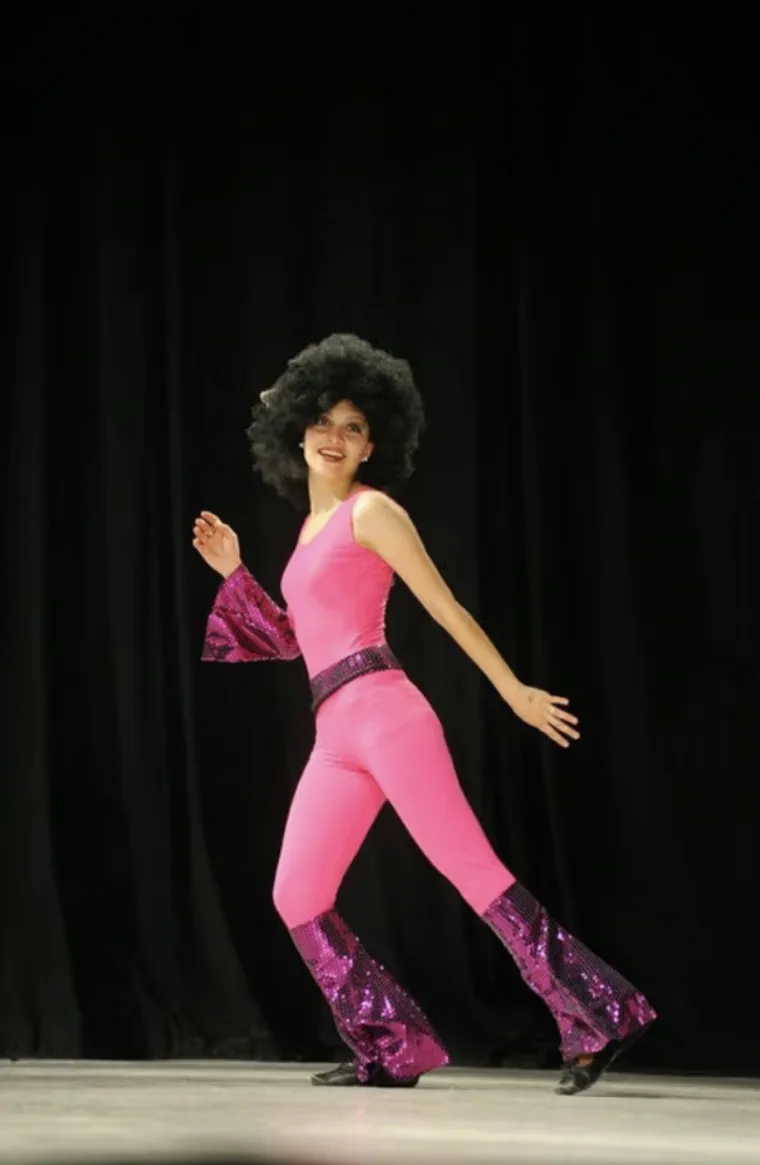 Una mujer con peluca afro negra y traje enterizo rosa baila sobre un escenario con fondo negro.