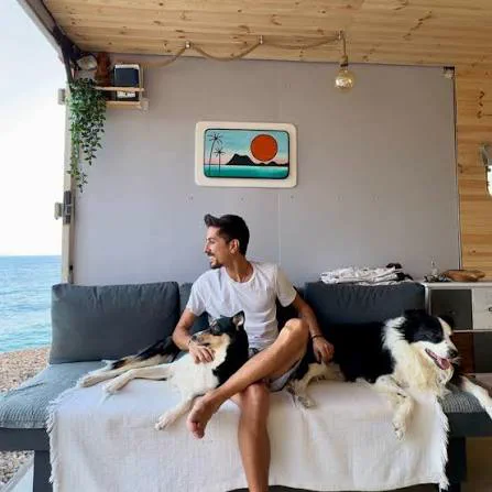 Un pibe sonríe sentado en un sillón junto al mar acompañado por dos perros.