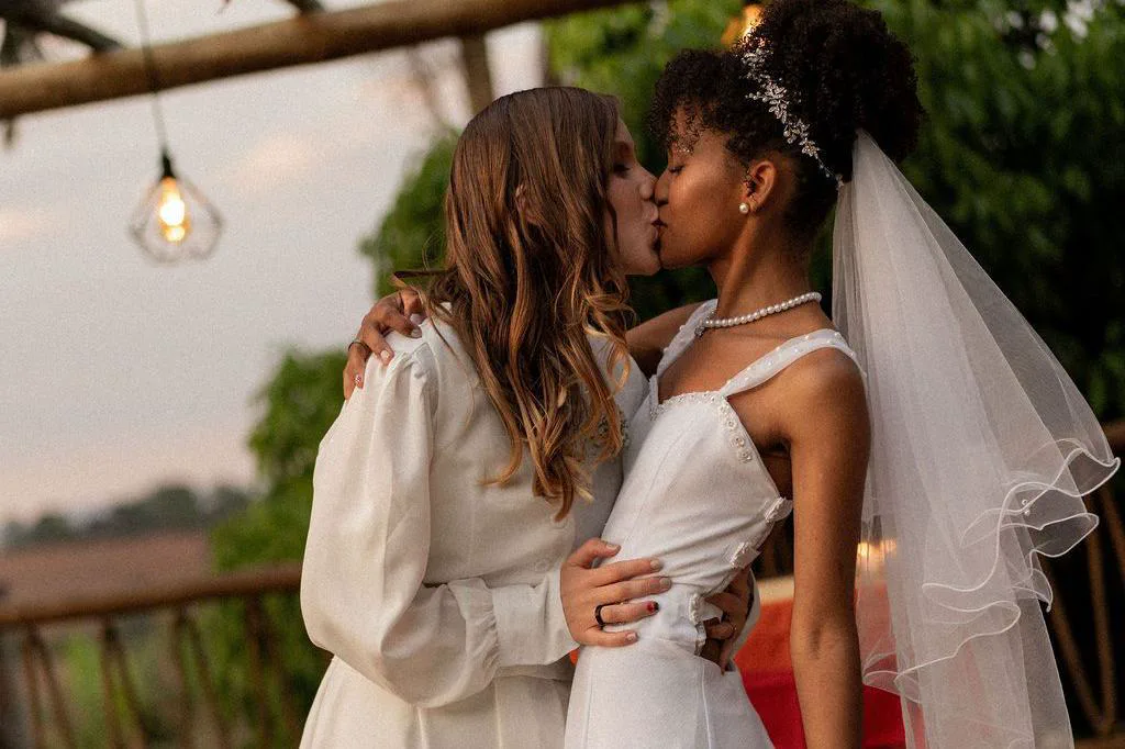 Dos mujeres vestidas de novia besándose al aire libre.