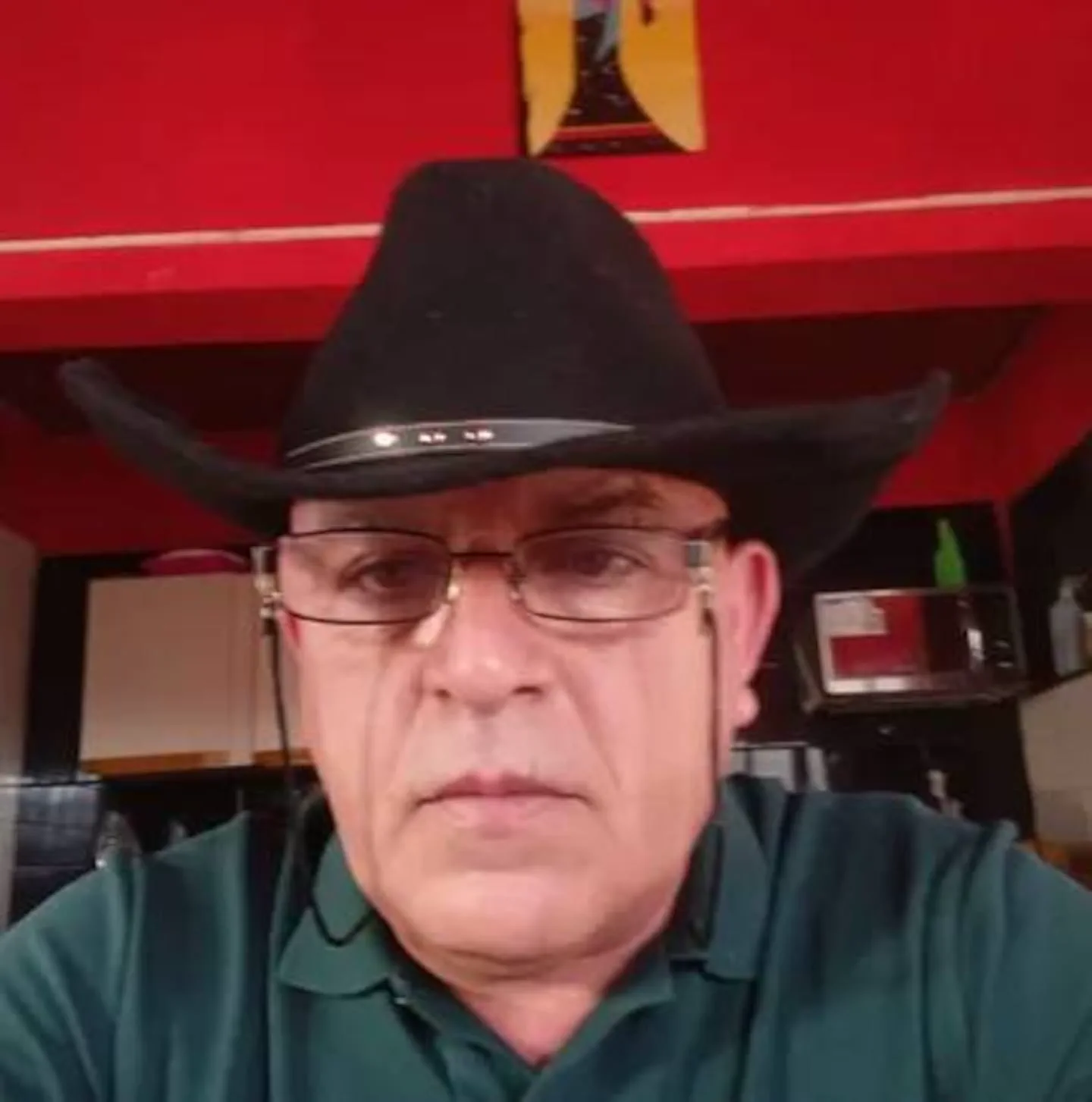 Un hombre con sombrero de vaquero negro, camisa verde y anteojos mira de frente hacia la cámara.