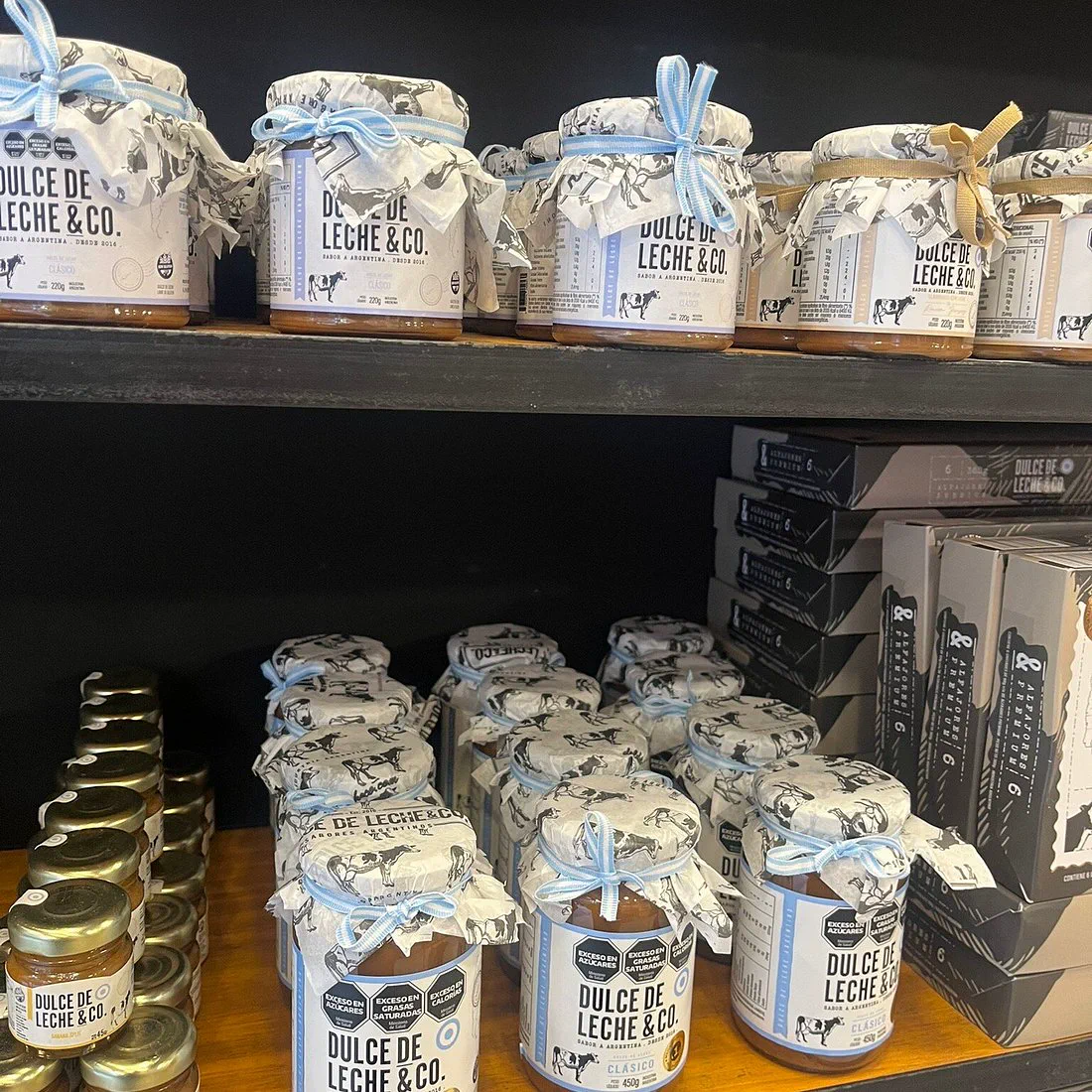 Varios frascos de dulce de leche y cajas de alfajores acomodados en estantes de madera oscura.