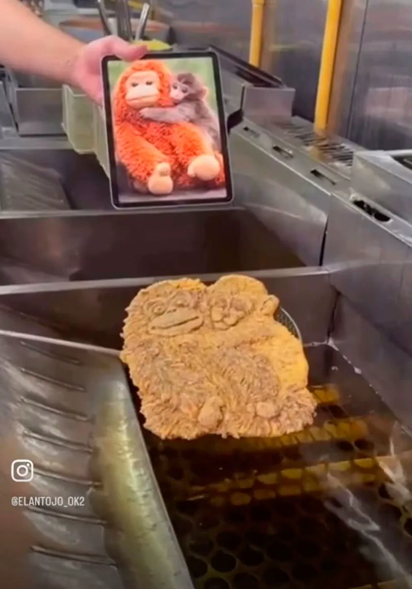 Una milanesa con forma de mono se fríe en una freidora mientras una persona sostiene una tableta con una foto.