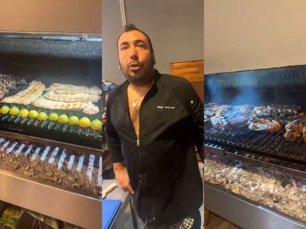 Un cocinero con chaquetilla negra está parado entre dos parrillas industriales con carne y limones al fuego.