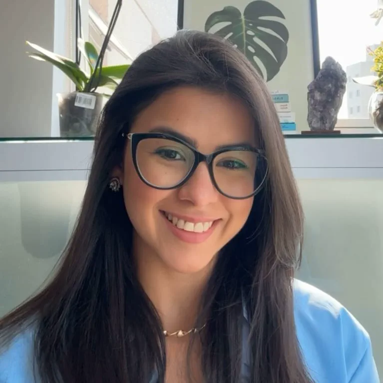 Una joven sonríe a cámara con anteojos negros, camisa celeste y plantas de fondo.