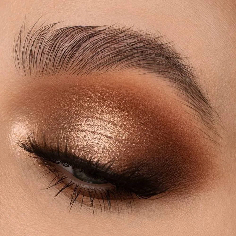 Primer plano de un ojo entrecerrado maquillado con sombra cobriza brillante y ceja peinada hacia arriba.