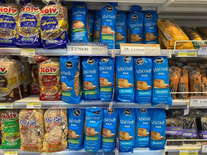 Paquetes de pan de molde y budines acomodados en los estantes de una góndola de supermercado con carteles de precios.