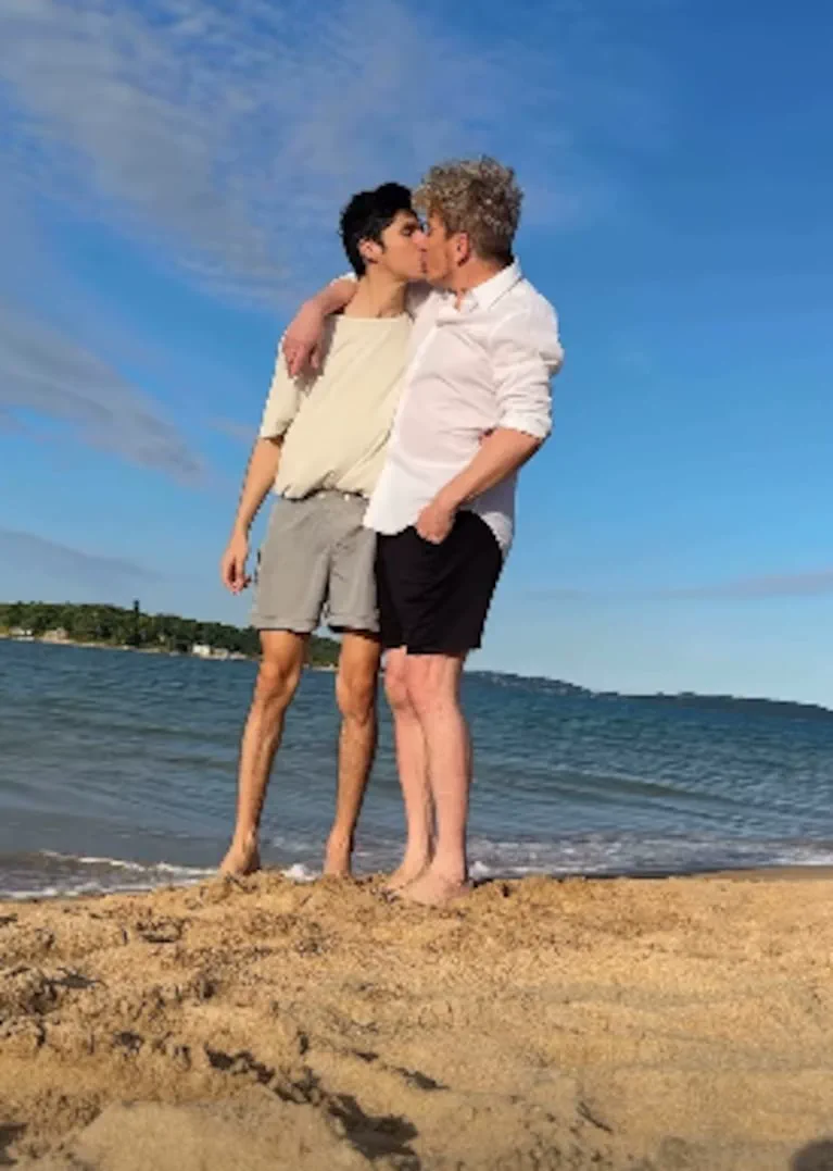 Dos hombres se abrazan y besan de pie sobre la arena de una playa frente al mar.