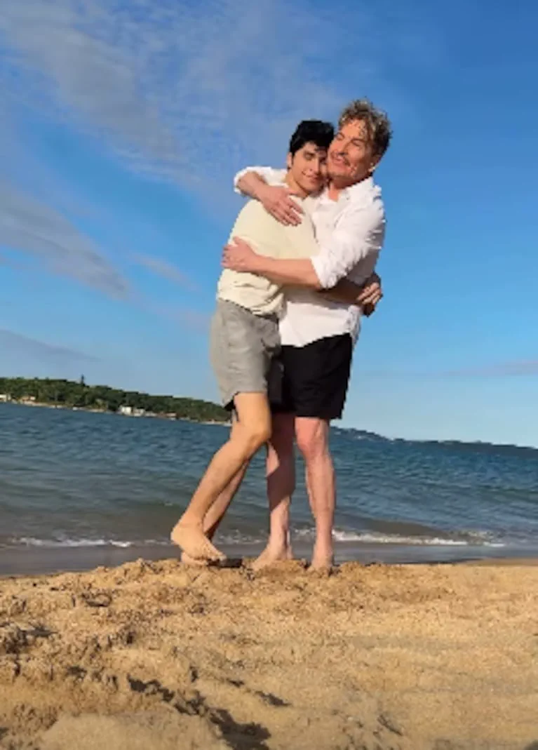 Dos hombres se abrazan descalzos en la arena de una playa frente al agua y bajo el cielo despejado.