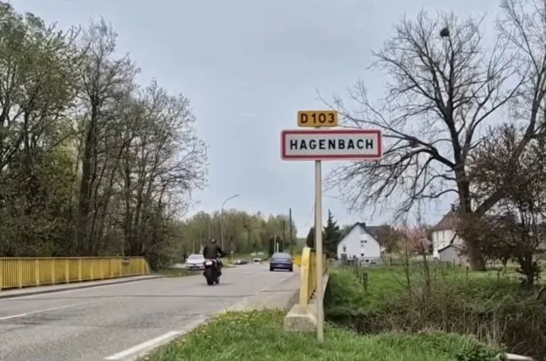 Cartel vial con el nombre Hagenbach al costado de una ruta con vehiculos circulando.