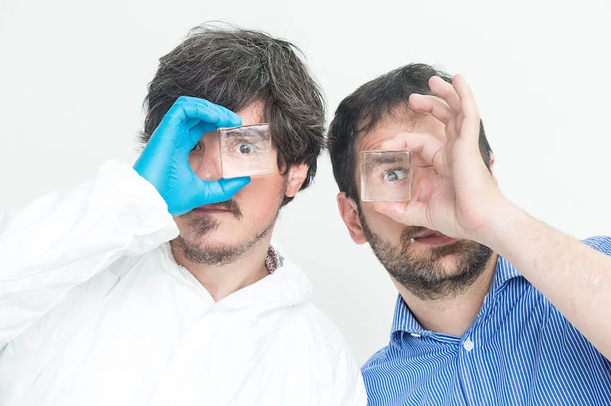 Dos hombres sostienen pequeños bloques transparentes frente a sus ojos sobre un fondo blanco.
