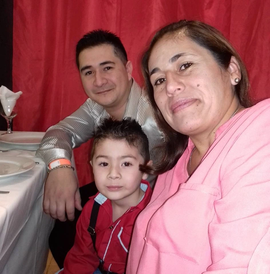 Un nene con camisa roja posa sentado entre dos adultos frente a una mesa y una cortina roja.