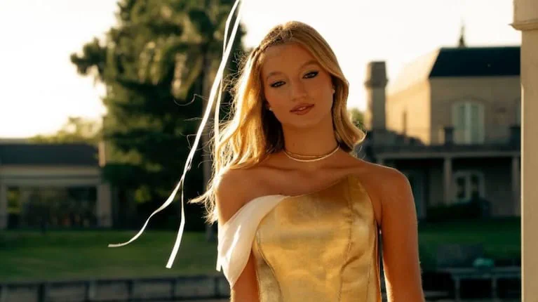 Indiana Cubero posa al aire libre con un vestido dorado frente a un parque y una casa.