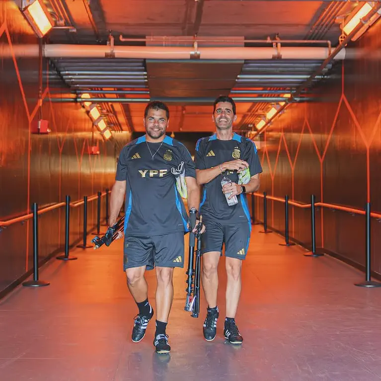 Dos hombres con indumentaria de la selección argentina caminan sonrientes por un pasillo iluminado.