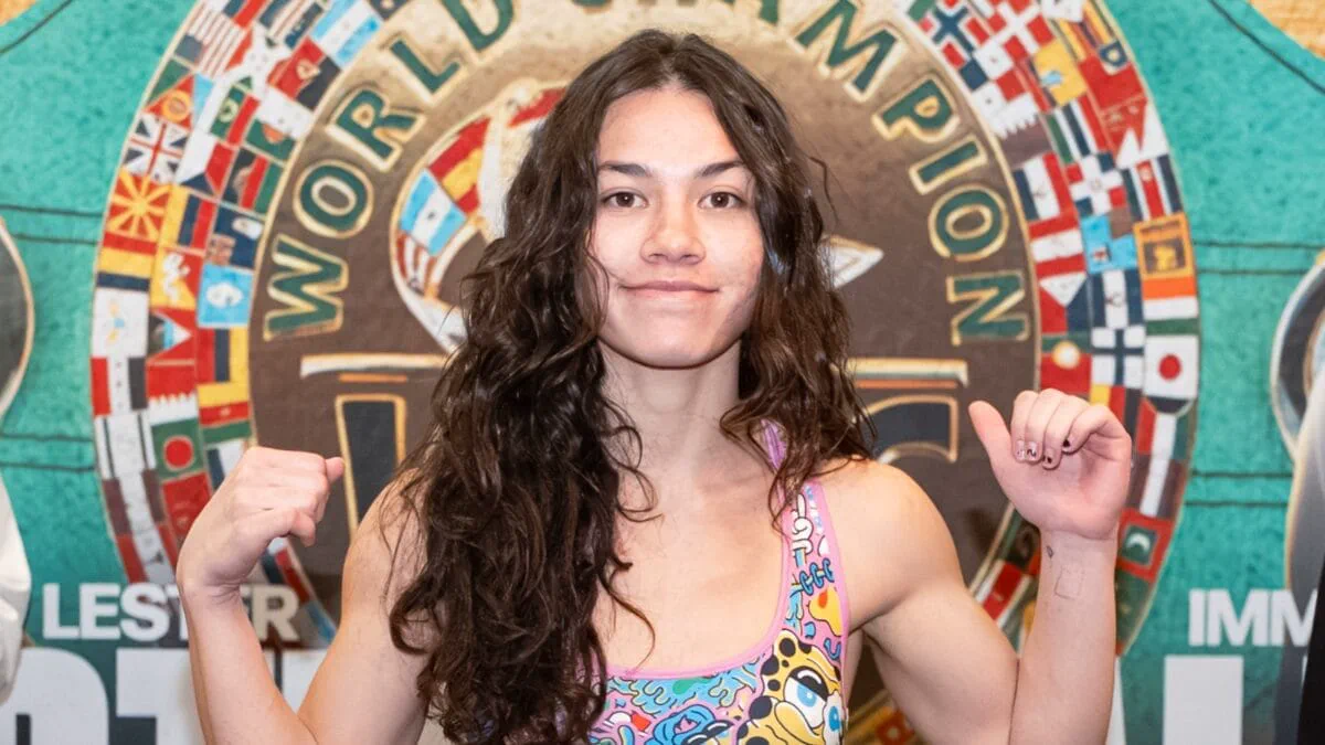 Una boxeadora de pelo largo ondulado posa levantando los brazos frente a un fondo con un cinturon de campeonato.