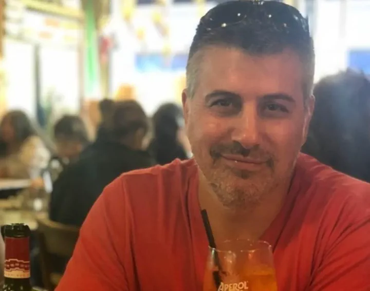 Un hombre de remera roja y barba candado sentado a una mesa con una copa con trago y una botella.