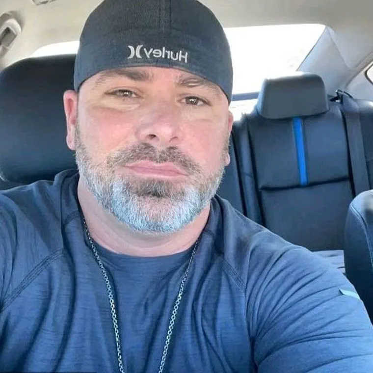 Un hombre con barba entrecana y gorra negra se saca una selfie en el interior de un vehiculo.