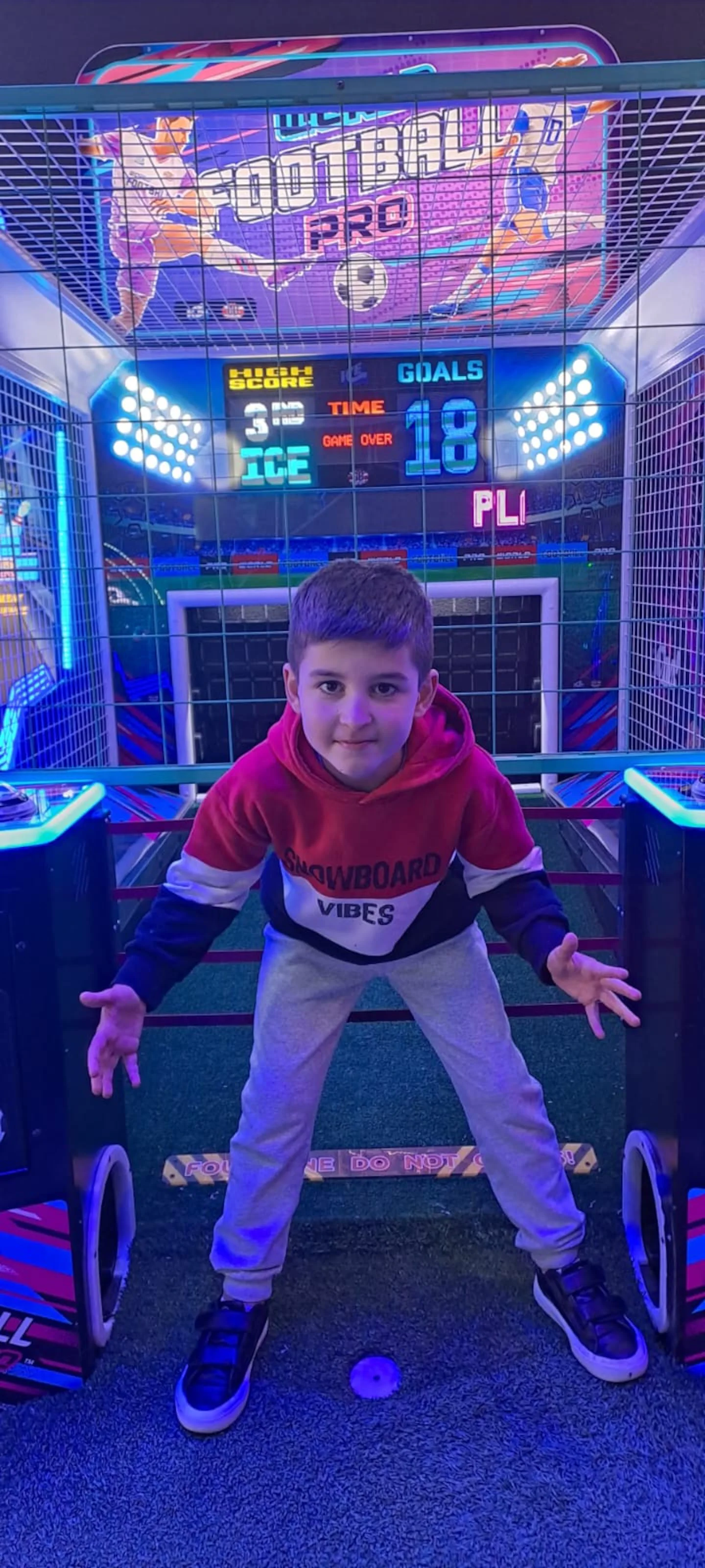 Un nene posa parado con los brazos abiertos delante de un juego arcade de fútbol.