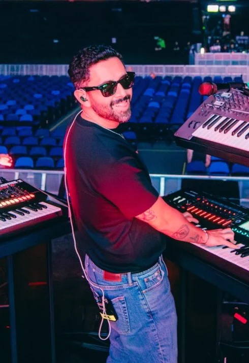 Un hombre con anteojos oscuros y remera negra tocando un teclado electrónico en un escenario.