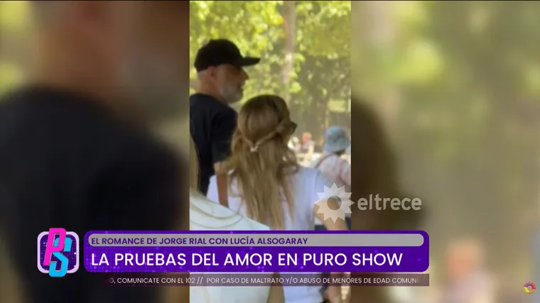 Jorge Rial camina de espaldas en un parque junto a una mujer con una remera blanca estampada.