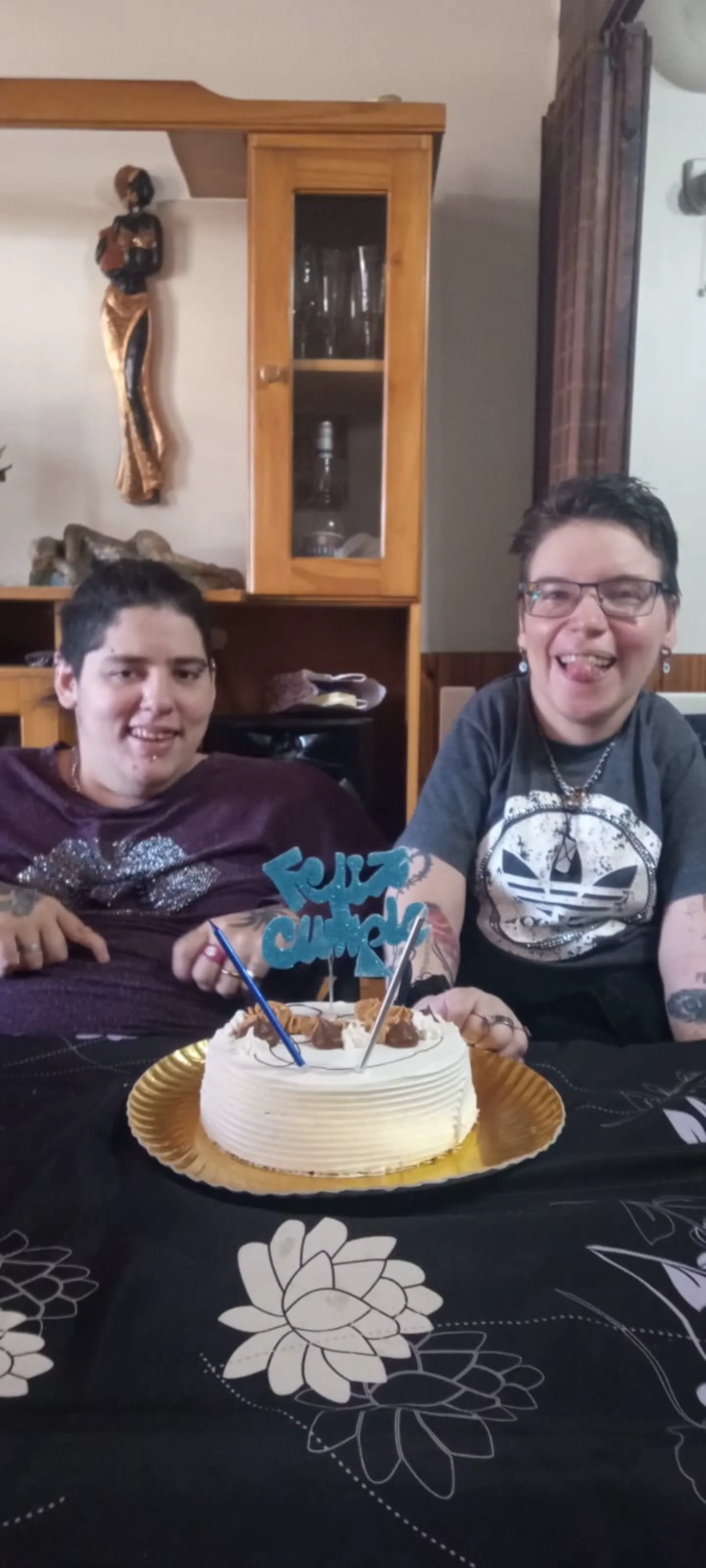 Dos personas sonríen sentadas a la mesa frente a una torta de cumpleaños con velas y adorno de feliz cumple.