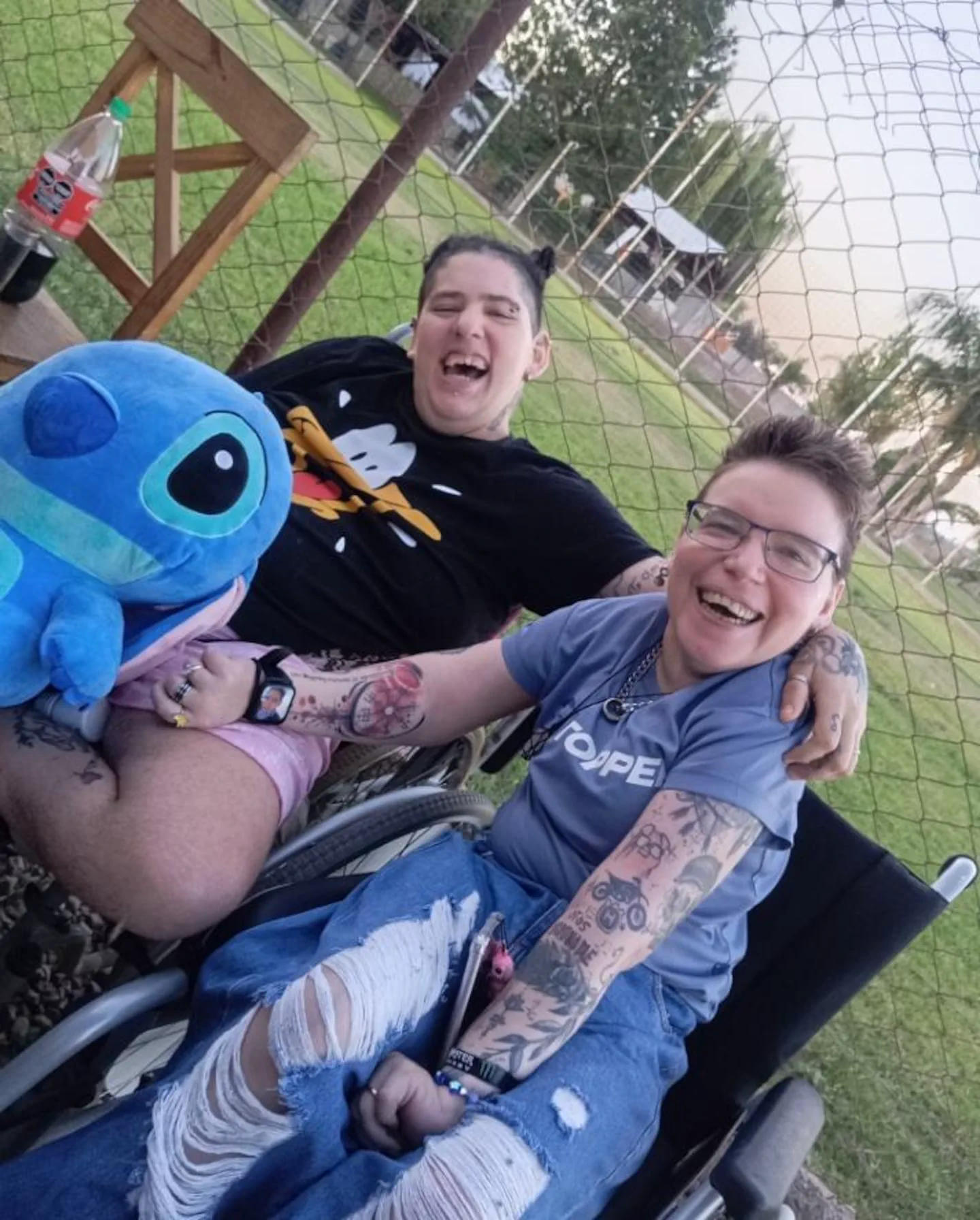 Dos personas sonríen al aire libre sentadas en sillas de ruedas, una de ellas sosteniendo un muñeco de peluche azul.