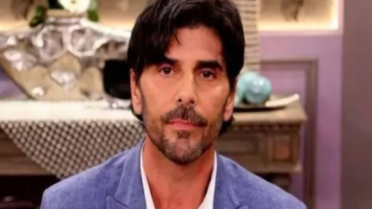 Un hombre con saco azul, camisa blanca y barba mira al frente en un interior.