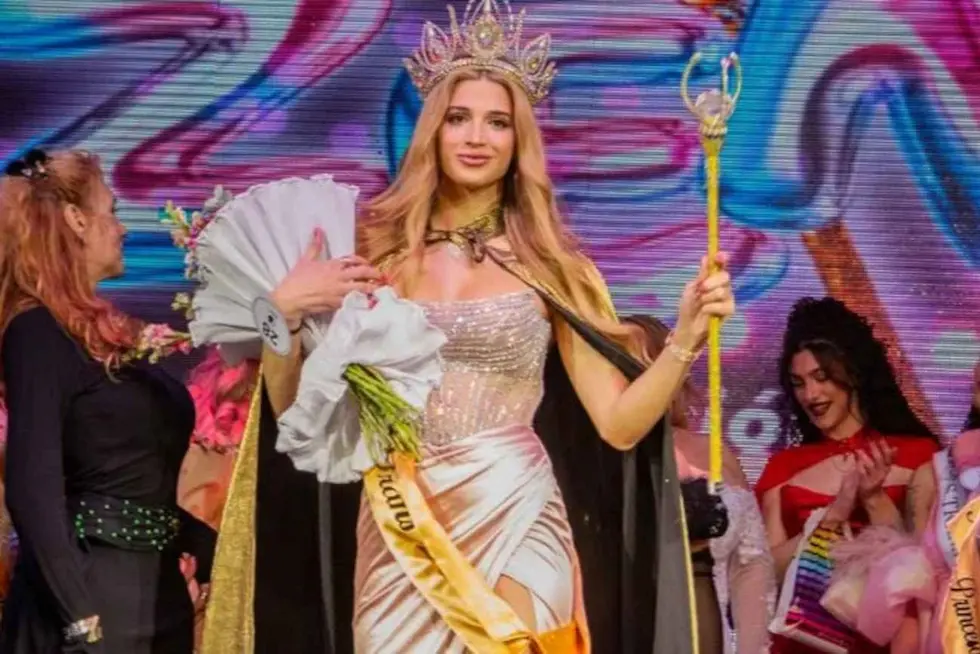 Una mujer rubia con corona y cetro sostiene un ramo de flores durante una premiación.