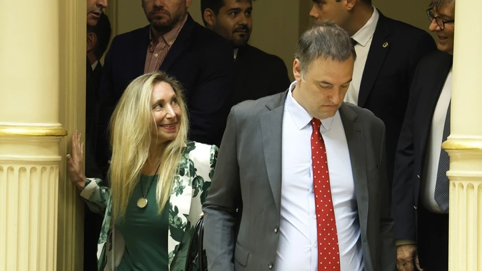 Karina Milei y Manuel Adorni caminan junto a otras personas de traje entre columnas.