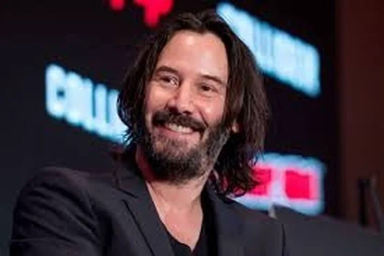 El actor Keanu Reeves sonriendo con saco negro y barba durante una presentacion.