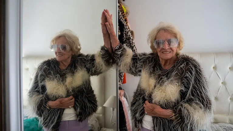 Mujer mayor con tapado de plumas y anteojos grandes apoyando la mano sobre un espejo.