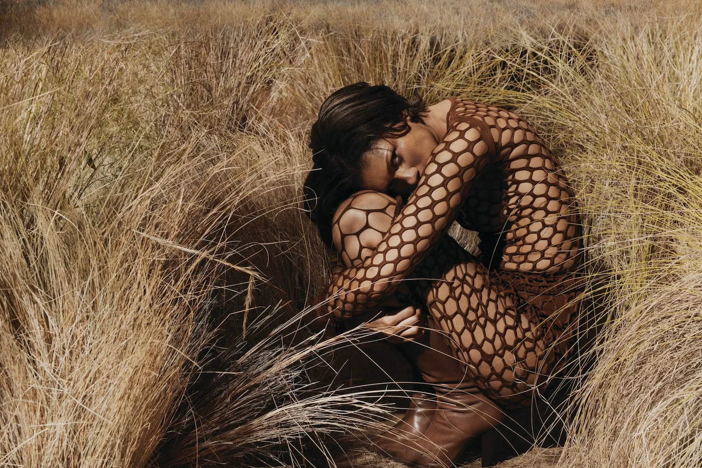 Zendaya aparece agachada entre pastizales secos vistiendo una prenda marrón calada de red y botas.