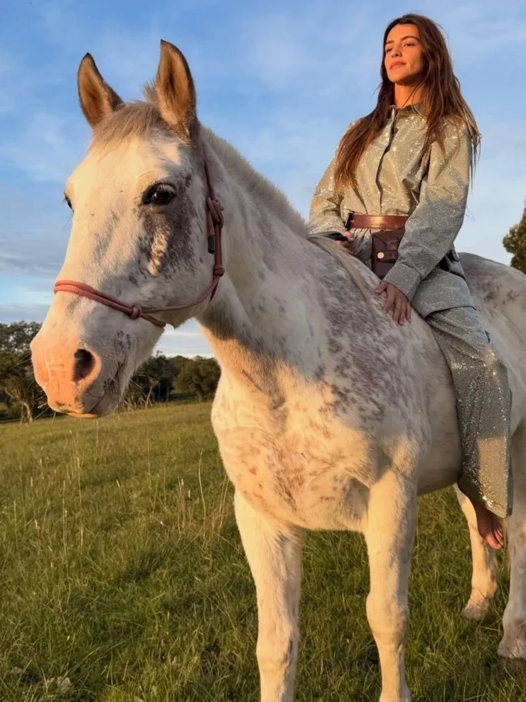 Una mujer con traje brilloso montando a caballo en un campo abierto bajo la luz del atardecer.